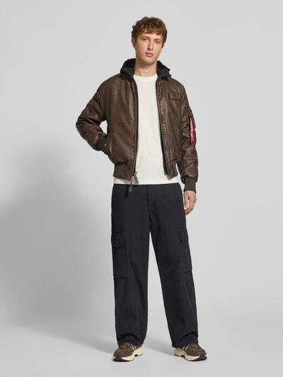 Männermodel trägt Bomberjacke in Leder-Optik mit Label-Details von Alpha Industries, dunkelbraun, Regular Fit, Ganzkörperansicht von vorne. Rückenlänge 63 cm, Schulterbreite 44 cm, Brustweite 52 cm, Ärmellänge 60 cm, Kapuze und Reißverschluss, strukturierte Oberfläche.