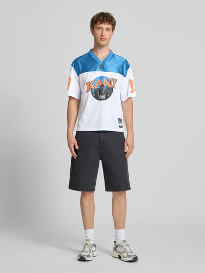Männermodel trägt Herren T-Shirt Trikot-Shirt mit Logo- und Motiv-Prints Modell 'OG Sport College' von KARL KANI im Boxy-Fit mit V-Ausschnitt, Label-Patch, Weiß und Blau, Rückenlänge 64 cm, Ganzkörperansicht von vorne.