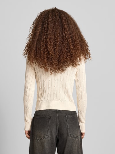 Frauenmodel trägt Strickpullover mit Zopfmuster und Label-Stitching von REVIEW, Baumwoll-Mix, Regular-Fit, Rundhalsausschnitt, Rückenlänge 52 cm, Ärmellänge 69 cm, cremefarben, feine Strickstruktur, dunkle Jeans, Ganzkörperansicht Rückansicht.