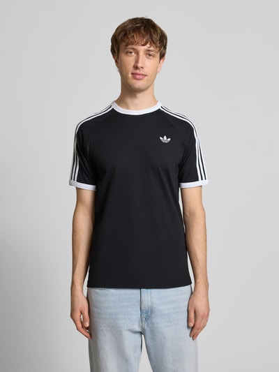 Männermodel trägt T-Shirt mit Label-Stitching von adidas Originals, schwarzer Baumwollstoff, weiße Details, Regular Fit, Ganzkörperansicht von vorne.