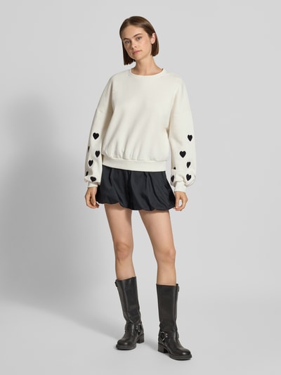 Frauenmodel trägt Regular Fit Sweatshirt aus Baumwoll-Mix Modell 'MACY' von ONLY, weiß mit schwarzen Herzmotiven, kombiniert mit schwarzem Rock und schwarzen Stiefeln, Damen Sweatshirt von ONLY, Ganzkörperansicht von vorne.