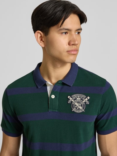 Männermodel trägt Slim Fit Poloshirt mit Label-Stitching und Knopfleiste von Superdry aus Reine Baumwolle, Nahaufnahme Oberkörper. Rückenlänge bei Größe M: 70 cm.