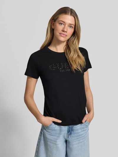 Frauenmodel trägt T-Shirt mit Label-Print und Strasssteinbesatz von Guess, schwarze Baumwolle, Straight Fit, Rundhals, Ganzkörperansicht von vorne
