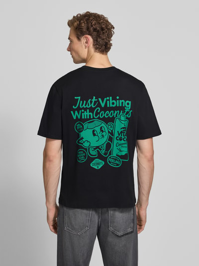 Männermodel trägt Regular Fit T-Shirt aus reiner Baumwolle Modell 'VIBING COCO' von REVIEW, Schwarz mit grünem Rückenprint, Ganzkörperansicht Rückansicht. Herren T-Shirt von REVIEW x ANSON´S, Rückenlänge bei Größe M: 68 cm, Print auf der Rückseite.