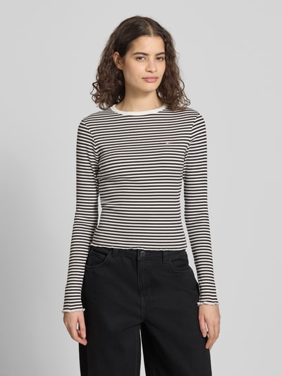 Kobiece model prezentuje Slim Fit Langarmshirt aus Baumwoll-Mix od Tommy Jeans, cienkie czarno-białe paski, logo na piersi, okrągły dekolt, długie rękawy z falbanką, delikatna prążkowana bawełna, krój slim fit, widok całej sylwetki z przodu.
