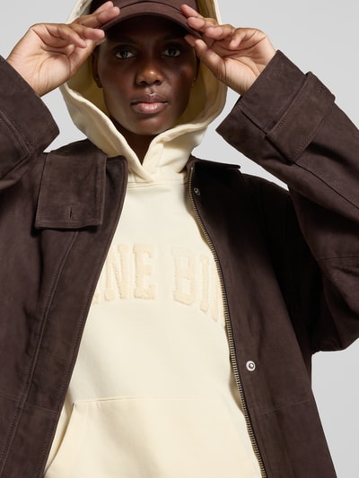 Frauenmodel trägt Oversized Hoodie aus reiner Baumwolle von Anine Bing in Creme mit Känguru-Tasche, Label-Detail und elastischen Abschlüssen, Nahaufnahme Oberkörper.