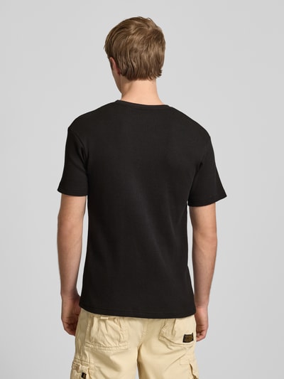 Männermodel trägt T-Shirt mit Label-Patch von Alpha Industries in Schwarz, Reine Baumwolle, Körpernahe Passform, Rundhalsausschnitt, Rückenlänge bei Größe M: 66 cm, Ganzkörperansicht Rückansicht.