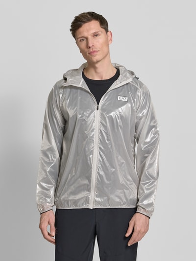 Männermodel trägt Blouson mit Label-Print von EA7 Emporio Armani in semitransparentem Grau, Kapuze, Reißverschluss, elastische Abschlüsse, Ganzkörperansicht von vorne.