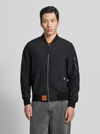 Männermodel trägt schwarze Bomberjacke mit Label-Patch und Eingrifftaschen von Original Bombers, Gerader-Schnitt, Ganzkörperansicht von vorne. Rückenlänge 67 cm, Schulterbreite 43 cm, Brustweite 53 cm, Ärmellänge 68 cm, Reißverschluss, Reißverschlusstasche am Ärmel, gerippte Abschlüsse, Druckknopfverschluss.