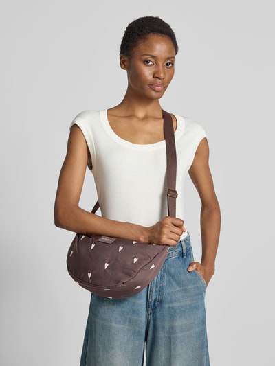 Kobiece model prezentuje Fanny Pack mit Logo-Detail Model 'Puffy' od Studio Noos, brązowy z wyhaftowanym logo na całej powierzchni, Główna przegroda z zamkiem błyskawicznym, regulowany pasek na ramię, szerokość w rozmiarze 1: 39 cm, długość w rozmiarze 1: 21 cm, głębokość w rozmiarze 1: 10 cm, długość paska na ramię w rozmiarze 1: 112 cm, detal z logo z przodu, widok całej sylwetki z przodu.
