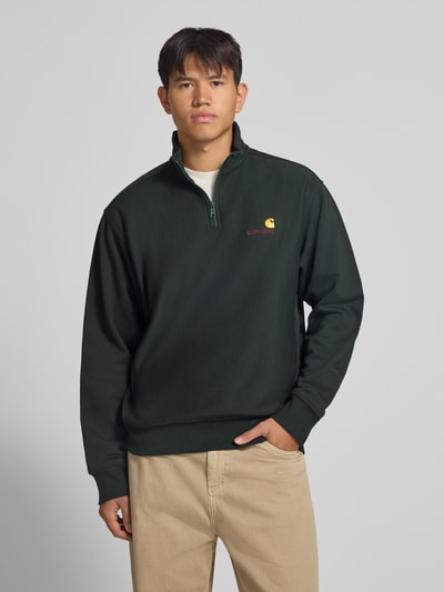 Męski model nosi Relaxed Fit Sweatshirt aus elastischem Material-Mix von Carhartt Work In Progress w kolorze ciemnej zieleni, haft z logo, elastyczne ściągacze, Troyer-kołnierz, materiał mieszany, długość tyłu 70 cm, długość rękawa 66 cm, widok całej sylwetki z przodu.