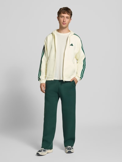 Männermodel trägt Regular Fit Sweatjacke aus Baumwoll-Mix von adidas in Creme mit grünen Streifen, Hose in Dunkelgrün, Ganzkörperansicht von vorne, Herren Sweatjacke von adidas Sportswear.