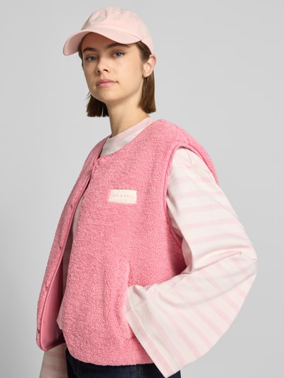 Kobiece model nosi Fleeceweste mit Druckknopfleiste Modell 'Magnolia' od OH APRIL, prosty krój, okrągły dekolt, listwa z zatrzaskami, naszywka z logo, wpuszczane kieszenie, długość tyłu w rozmiarze S: 51 cm, szerokość ramion w rozmiarze S: 45 cm, szerokość klatki piersiowej w rozmiarze S: 60 cm, różowa faktura frotte, widoczne białe i różowe paski, ujęcie z przodu, zbliżenie na górną część ciała.