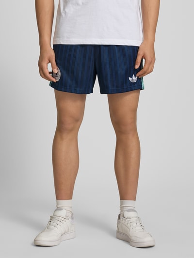 Mężczyzna nosi Loose Fit Trikot-Shorts mit DFB-Applikation od adidas Originals w granatowym kolorze z wzorem allover, logo, kontrastowymi paskami, elastycznym ściągaczem i bocznymi kieszeniami, tkanina typu żakard, widok na całe nogi, ujęcie od przodu.