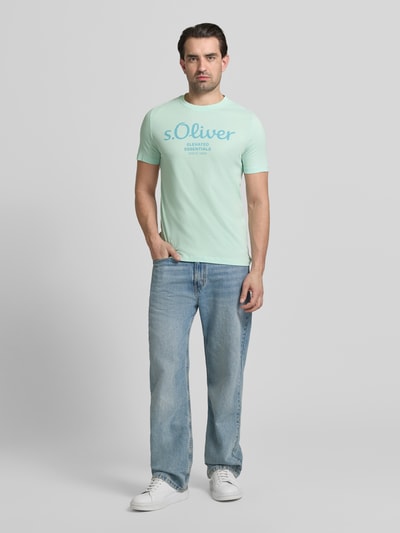 Męski model nosi Regular Fit T-Shirt aus reiner Baumwolle von s.Oliver RED LABEL, jednolita optyka, krój regular fit, nadruk z logo, okrągły dekolt, krótki rękaw, jersey z czystej bawełny, jasne kolory, gładka tekstura, całopostaciowy widok z przodu.