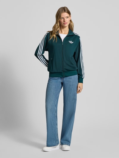 Frauenmodel trägt bluza rozpinana z stójką od adidas Originals, luźny krój, zamek błyskawiczny, wyhaftowane logo, kieszenie zapinane na zamek błyskawiczny, prążkowane wykończenia, ciemnozielona z białymi detalami, gładka tekstura, jeansy, białe buty, całkowity widok z przodu.