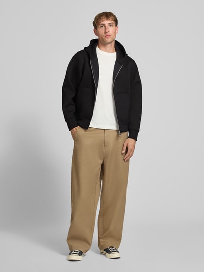 Männermodel trägt Relaxed Fit Sweatjacke mit Zweiwege-Reißverschluss von JACK & JONES in Schwarz mit Känguru-Tasche, elastischer Material-Mix, Ganzkörperansicht von vorne.