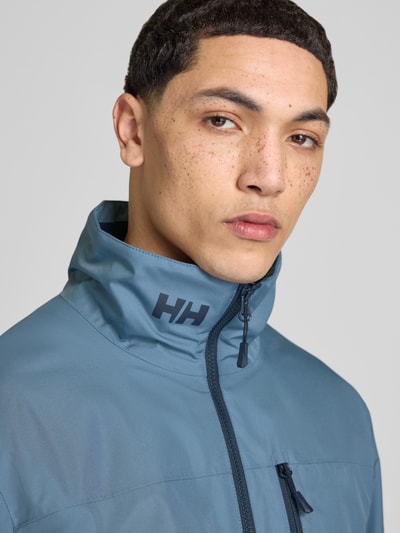 Męski model nosi Midlayer Jacke mit Stehkragen Modell 'CREW' od Helly Hansen, prosty krój, stójka, nadruk z logo, zamek błyskawiczny, brustasza zapinana na zamek błyskawiczny, naszywka z logo, kieszenie zapinane na zamek błyskawiczny, niebieski, tekstura gładka, nahaufnahme górnej części ciała.