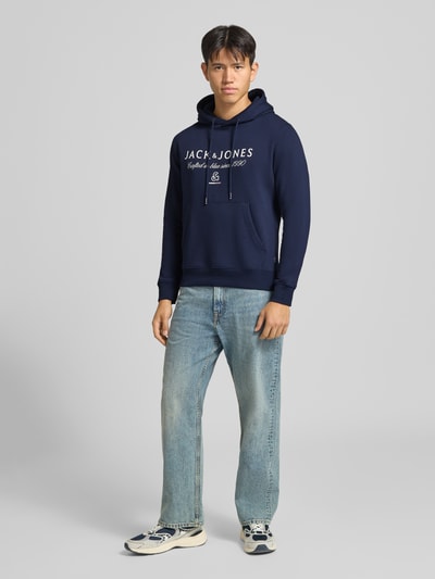 Männermodel trägt dunkelblauen Relaxed Fit Hoodie aus Baumwoll-Mix Modell 'KYLE' von JACK & JONES mit Label-Detail, Ganzkörperansicht von vorne.