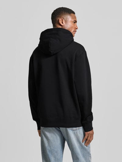 Männermodel trägt schwarzen Hoodie mit Logo-Print und Känguru-Tasche von Wood Wood, reine Baumwolle, lockere Passform, Ganzkörperansicht Rückansicht.