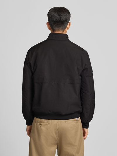 Männermodel trägt Regular fit Heren jack Model 'HARRINGTON' von Superdry aus elastische materiaalmix in zwart, gestikt logo, opstaande kraag, ritssluiting, ruglengte maat M 66 cm, schouderbreedte 50 cm, borstwijdte 60 cm, mouwlengte 65 cm, ganzkörperansicht rückansicht.