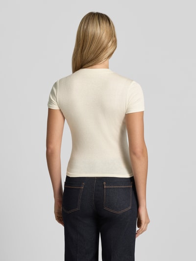 Vrouwenmodel draagt gebreid shirt van viscosemix Model 'LICIA' van GUESS in licht beige, slim fit, gladde structuur, geheel aanzicht achterkant. Dames gebreid shirt, viscosemix, slim fit, logo met siersteentjes, ronde hals, ruglengte bij maat S 52 cm.
