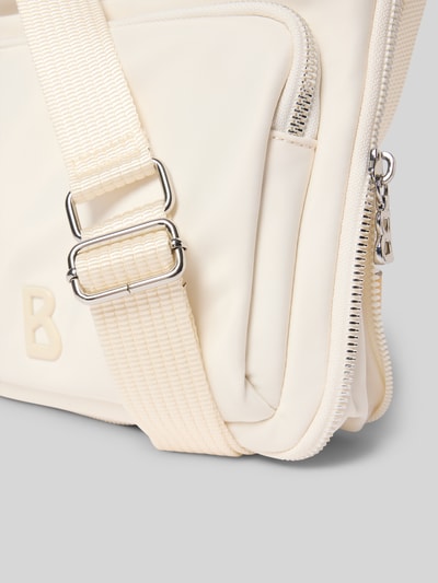 Produktfoto, cremefarbene Handtasche mit Label-Detail Modell 'verbier play 1.0 serena' von BOGNER, Reißverschluss, verstellbarer Schulterriemen, glatte Textur, Detailansicht. Breite 25 cm, Länge 25 cm, Tiefe 4 cm, Riemenlänge 105 cm.