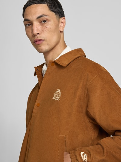 Männermodel trägt Loose Fit Bomberjacke aus reiner Baumwolle von Nike in Braun mit Logo-Details, Nahaufnahme Oberkörper. Herren Bomberjacke / Blouson