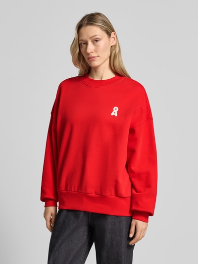 Frauenmodel trägt Oversized Sweatshirt aus reiner Baumwolle Modell 'ICONIC A ALIZAA' von Armedangels in Unifarbe Rot mit Logo-Stitching, Struktur: weiches Sweatmaterial, Crew Neck, gerippte Abschlüsse, Ganzkörperansicht von vorne.
