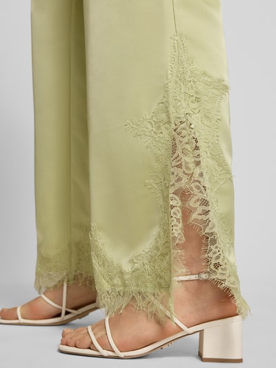 Frauenmodel trägt Flared Fit Stoffhose mit Spitzenbesatz von Gina Tricot in Satin-Optik, grün, Spitzenbesatz am Bein, Nahaufnahme Bein. Damen Stoffhose, elastischer Bund, Tunnelzug, Bundweite 40 cm, Innenbeinlänge 79 cm, Größe S.