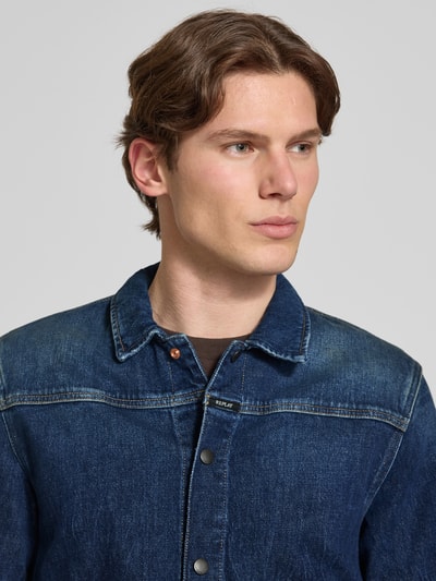 Mężczyznamodel nosi Jeansjacke mit Pattentaschen und Kentkragen od Replay, mieszanka bawełny z dodatkiem streczu, zapięcie na zatrzask, detal z logo, ciemnoniebieski denim, widoczne kieszenie z patką, krój straight fit, kołnierzyk typu kent, szerokość w rozmiarze M: 51 cm, długość tyłu: 68 cm, szerokość ramion: 38 cm, długość rękawów: 66 cm, tekstura gładka, lekko sprany efekt, nahaufnahme górnej części ciała.