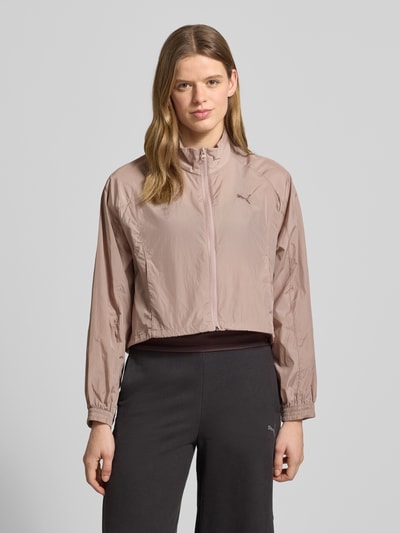 Frauenmodel trägt Relaxed Fit Jacke mit Stehkragen Modell 'Her' von PUMA in Beige mit Logo-Stitching, Reißverschluss und Eingrifftaschen, elastische Ärmelabschlüsse, Ganzkörperansicht von vorne. Rückenlänge bei Größe S: 55 cm, Schulterbreite: 45 cm, Brustweite: 56 cm, Ärmellänge: 64 cm.