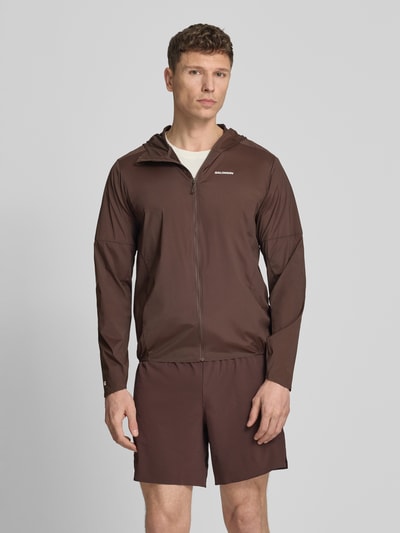 Männermodel trägt Windbreaker mit Kapuze und Logo Modell 'SENSE AERO' von Salomon aus Material-Mix mit Stretch-Anteil, Regular-Fit, Reißverschluss, Rückenlänge 71 cm, Schulterbreite 44 cm, Brustweite 41 cm, Ärmellänge 67 cm in Braun, Ganzkörperansicht von vorne.