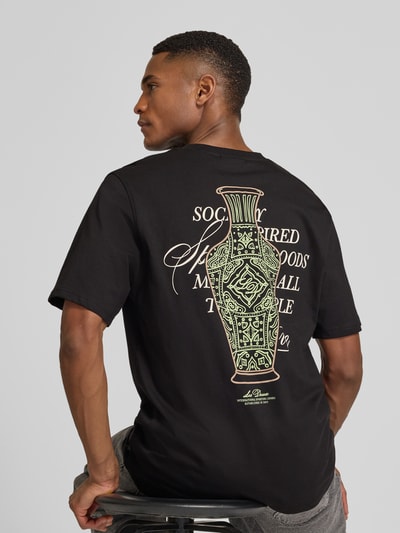 Männermodel trägt T-Shirt mit rückseitigem Motiv-Print Modell 'Ben Faience' von Les Deux, reine Baumwolle, krój regular fit, schwarz mit Statement-Print und Label-Detail, Ganzkörperansicht Rückansicht