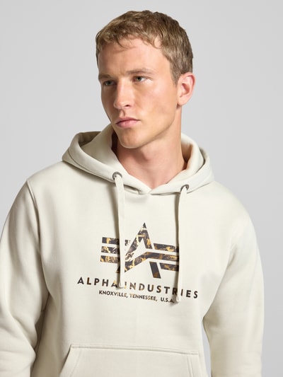 Männermodel trägt Hoodie mit Logo und Känguru-Tasche von Alpha Industries im Baumwoll-Mix, Regular Fit, cremefarben, Nahaufnahme Oberkörper.