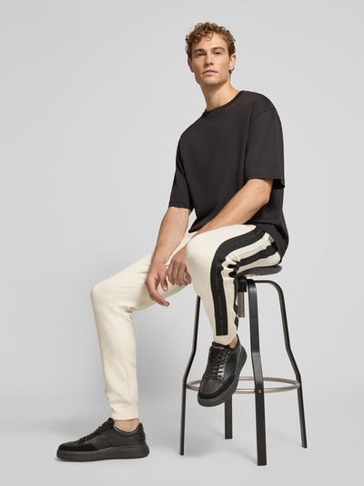 Männermodel trägt Sweatpants mit Bordüre von Armani Exchange in Beige mit schwarzem Label-Print und geradem Schnitt, Nahaufnahme Oberkörper, Herren Sweatpants, elastischer Baumwoll-Mix, Elastikbund mit Tunnelzug, seitliche Eingrifftaschen, Gesäßtasche, Innenbeinlänge 79 cm bei Größe M.