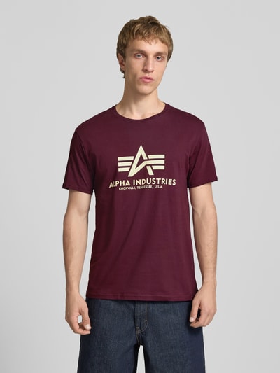 Męski model nosi T-Shirt mit Logo und Rundhalsausschnitt od Alpha Industries, bawełna, krój regular fit, kolor bordowy, widok całej sylwetki z przodu.