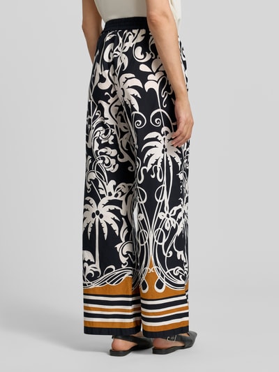 Frauenmodel trägt Wide Fit Stoffhose mit floralem Allover-Print von Emily Van den Bergh, schwarze und cremefarbene Muster, elastischer Bund, reine Baumwolle, lockerer Schnitt, Bundweite 105 cm, Innenbeinlänge 77 cm, orange-schwarze Streifen am Saum, Ganzkörperansicht Rückansicht.
