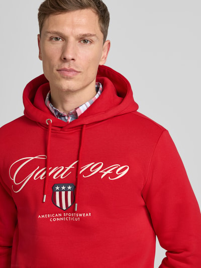 Męski model nosi czerwony Hoodie mit Label-Stitching und Känguru-Tasche von Gant, bawełniany, regular fit, zbliżenie na górną część ciała.