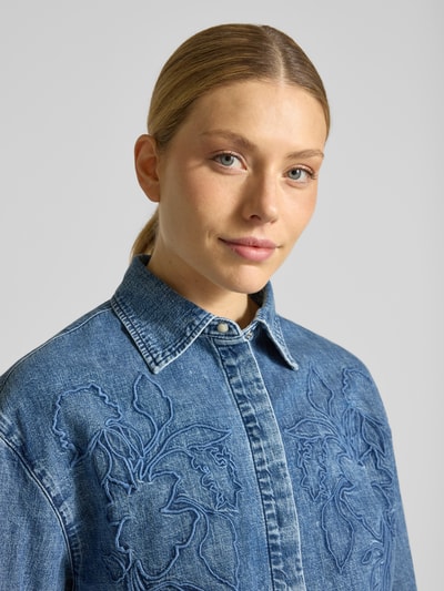 Modelka nosi Loose Fit Jeansbluse aus reiner Baumwolle von s.Oliver RED LABEL, tkanina stylizowana na denim, splot twill, haftowany motyw, długi rękaw, kołnierzyk koszulowy, napis, czysta bawełna, zapięcie na ukrytą listwę guzikową, niebieski odcień, dekolt w serek, detal na plecach, kontrafałda, zbliżenie na górną część ciała.
