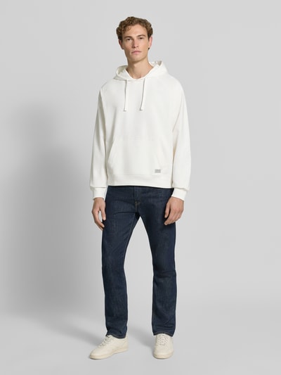 Męski model nosi Relaxed Fit Hoodie aus Baumwoll-Mix mit Logo-Patch od GUESS, biały, mieszanka bawełny, naszywka z logo, kaptur z tunelem, kieszeń kangurka, długość tyłu w rozmiarze M 70 cm, długość rękawów w rozmiarze M 79 cm, pełen widok z przodu
