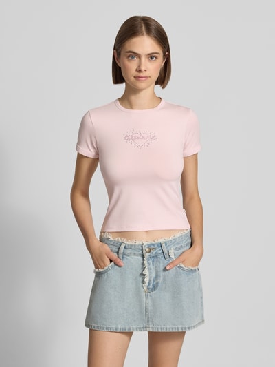 Frauenmodel trägt Slim Fit T-Shirt mit Strasssteinbesatz Modell 'Heart Baby' von GUESS Jeans in Rosa, Rundhalsausschnitt, Ganzkörperansicht von vorne.