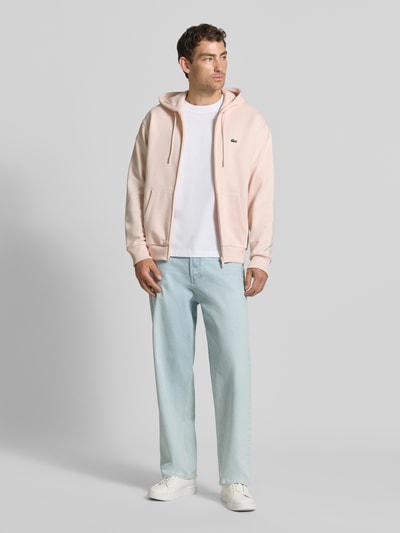 Männermodel trägt Regular Fit Sweatjacke aus Baumwoll-Mix von Lacoste in Hellrosa, kombiniert mit heller Jeans und weißen Sneakers, Ganzkörperansicht von vorne. Herren Sweatjacke von Lacoste.