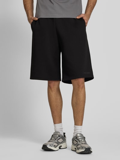 Männermodel trägt schwarze Sweatshorts mit Logo-Print von REVIEW, Material-Mix mit Baumwoll-Anteil, Loose-Fit, Ganzkörperansicht von vorne