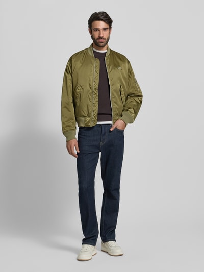 Mannelijk model draagt Regular Fit Bomberjack uit satijn van Lacoste in effen olijfgroen met logopatch, waterafstotend, warmte-isolatie, zijpaspelzakken, collegekraag en geribde boorden, geheel te zien van voren.