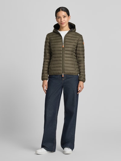 Frauenmodel trägt olivgrüne Steppjacke mit Zweiwege-Reißverschluss Modell 'DAISY' von Save The Duck, glatte Textur, Ganzkörperansicht von vorne. Kontext: Damen Steppjacke, Slim Fit, Kapuze, Reißverschlusstaschen, Rückenlänge 57 cm, Schulterbreite 44 cm, Brustweite 48 cm, Ärmellänge 60 cm.