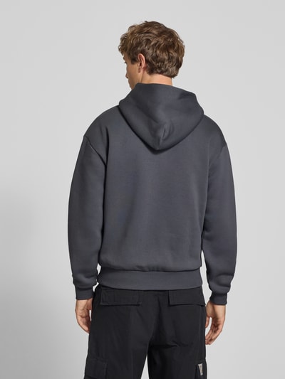 Mannenmodel draagt Relaxed Fit Sweatshirt aus Baumwoll-Mix mit Känguru-Tasche van REVIEW in donkergrijs met labeldetail, katoenmix, relaxed fit, capuchon, elastische boorden, ruglengte maat M 68 cm, mouwlengte maat M 62 cm, kangoeroezak, textuur glad, geheel rugaanzicht.