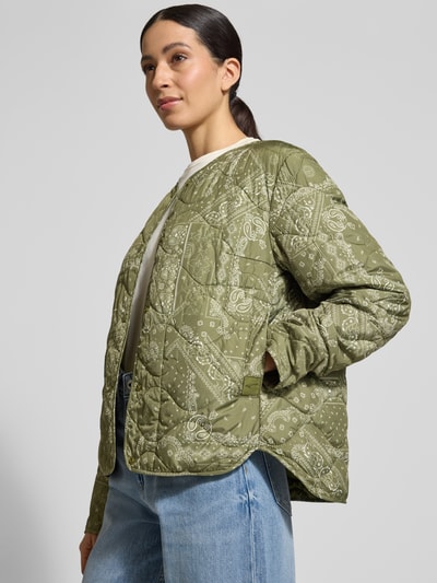 Modelka kobieca nosi Steppjacke mit Rundhalsausschnitt od camel active w kolorze oliwkowym z białym wzorem paisley, krój regular fit, pikowany materiał, zapięcie na napy, prosty fason, Szerokość w rozmiarze 36: 53 cm, długość tyłu: 60 cm, szerokość ramion: 43 cm, długość rękawa: 64 cm, widok całej sylwetki z przodu.