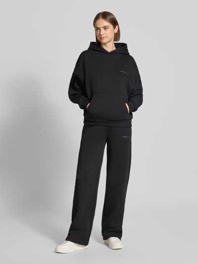 Frauenmodel trägt Hoodie mit Logo-Print und Känguru-Tasche von REVIEW, Schwarz, lockerer Baumwoll-Mix, Ganzkörperansicht von vorne.