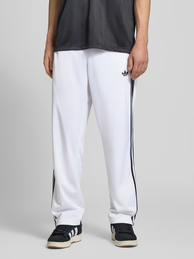 Männermodel trägt Regular Fit Sweatpants mit Label Print von adidas Originals in wit met all-over motief, elastische boord en zij-steekzakken, ganzkörperansicht von vorne.
